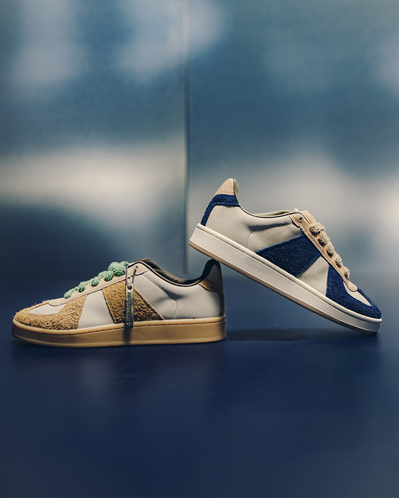 Two stylish low-top streetwear sneakers with suede and leather panels in tan and blue tones on a reflective blue surface.