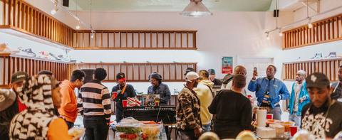 Group of people socializing at a men's streetwear sneaker shop event with food and a DJ.