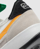 Close-up of sneaker heel with beige suede, black and white detail, orange stripe, and green pull tab