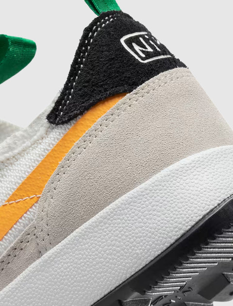 Close-up of sneaker heel with beige suede, black and white detail, orange stripe, and green pull tab