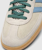 Close-up of suede sneaker with green laces, blue stripes, and gum sole.